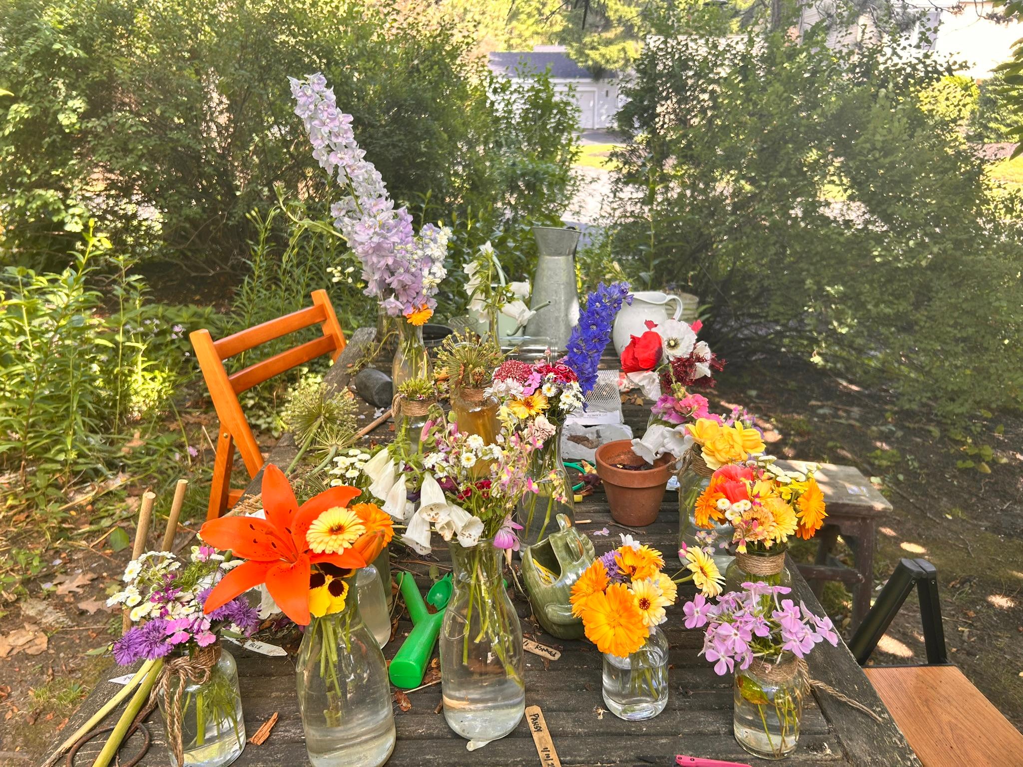 Garden worktable covered with small bouquets, jars, lilies, and flower supplies.