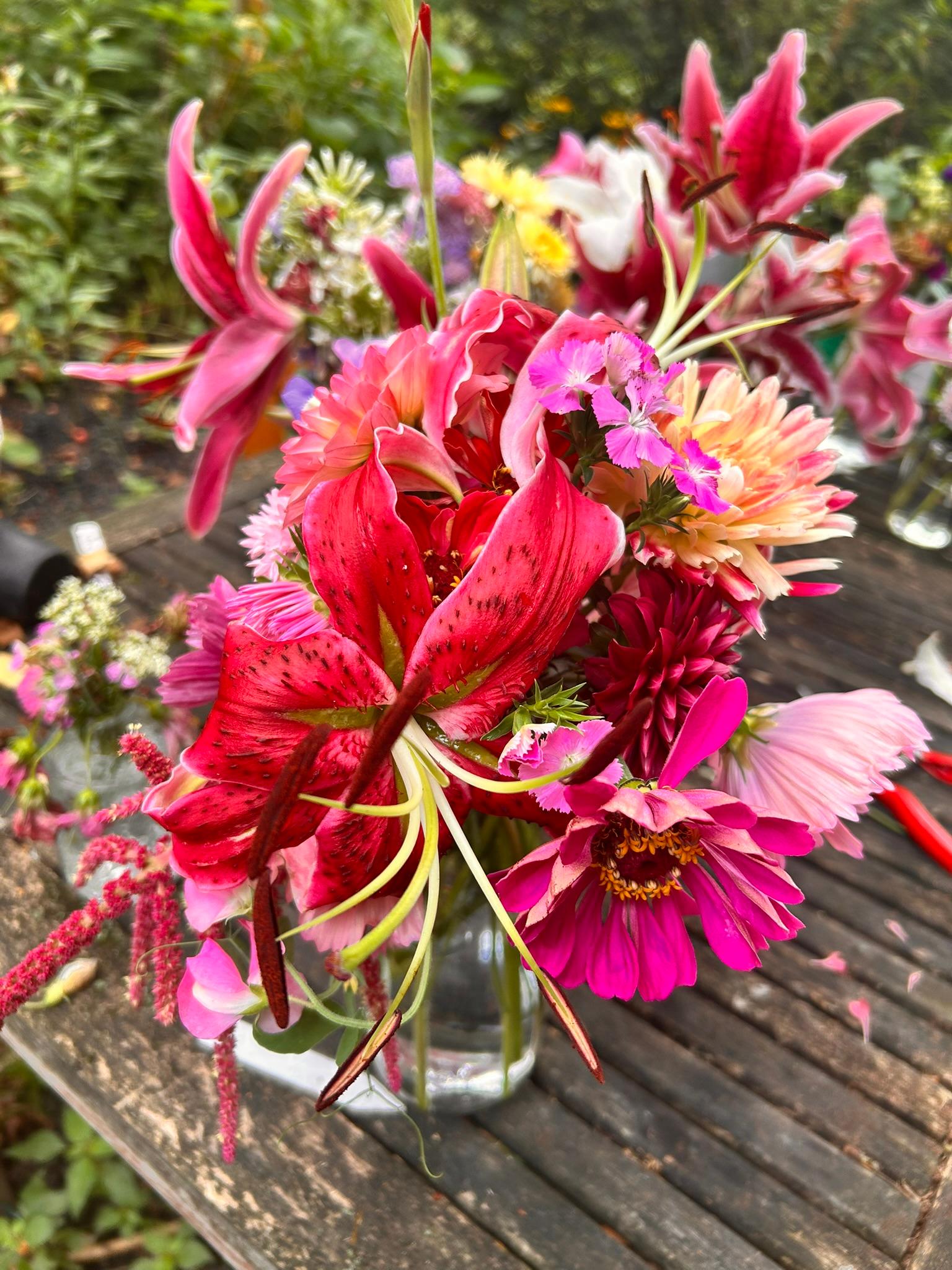 Bright local bouquet with pink lilies, dahlia, lisianthus, scabiosa, amaranth, and cosmos arranged on a weathered garden table.