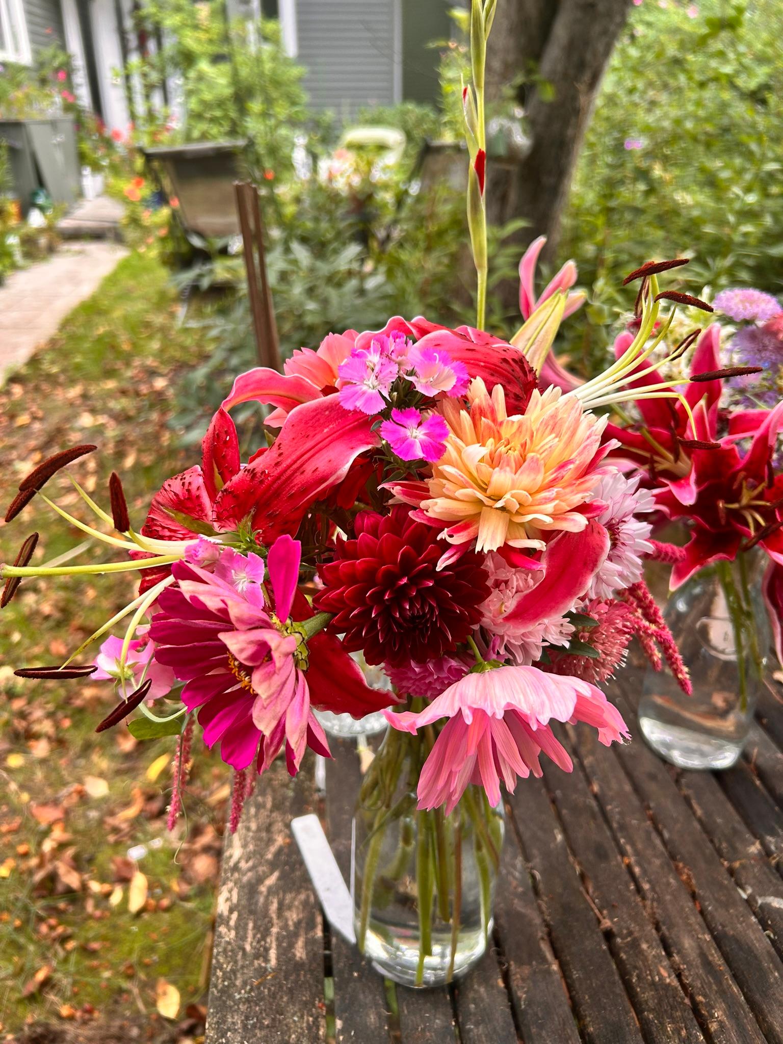 Bold pink bouquet with lilies and dahlias arranged in a glass vase on a weathered garden table.